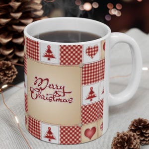Peut inclure: Un mug en céramique blanche orné d'un motif patchwork composé de carrés vichy rouges et blancs, de cœurs rouges et d'illustrations de sapins de Noël rouges. Le texte Merry Christmas est inscrit en rouge et en cursive sur fond beige.