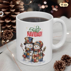 May include: A white 11 oz ceramic mug featuring a graphic of two snowmen wearing patchwork hats and scarves. The text "Feliz NAVIDAD!" is printed in green and red above the snowmen. The snowmen are decorated with small twigs and a heart.