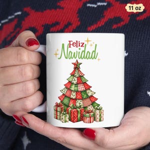 May include: A white 11 oz ceramic mug featuring a graphic of a patchwork-style Christmas tree in red and green patterns, surrounded by wrapped gift boxes at the base. The text "Feliz Navidad" is printed in red and green above the tree.