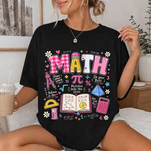 May include: A black t-shirt featuring a colorful graphic with the word MATH in large letters, surrounded by various mathematical symbols, a calculator, a protractor, a compass, and an open textbook with equations and the text Mistakes Help You Grow.