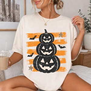 May include: A cream-colored short-sleeve t-shirt featuring a graphic of three stacked black jack-o'-lanterns centered over horizontal orange brushstrokes. The design includes small black bats and grey stars scattered around the pumpkins.