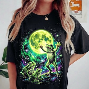 May include: A black short-sleeve t-shirt featuring a graphic print of three green frogs in a cosmic setting. One frog stands with arms raised toward a large, glowing yellow moon, while two others sit below amidst vibrant green and purple nebula clouds.