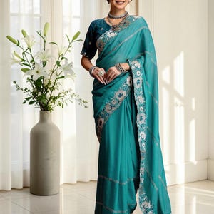 Op de afbeelding: Een blauwgroene sari met een bijpassende blouse met korte mouwen. Het kledingstuk is versierd met ingewikkeld zilverkleurig bloemenborduurwerk langs de randen en horizontale banden. De stof heeft een gladde textuur en een gedrapeerde pallu.