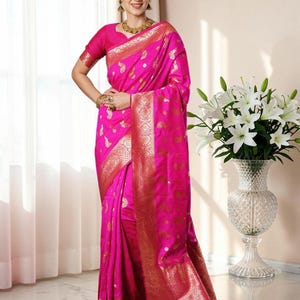 May include: A vibrant fuchsia pink saree featuring intricate gold zari woven patterns throughout the fabric. The garment includes a wide, ornate gold border with traditional motifs and a matching short-sleeved blouse, creating a cohesive look.