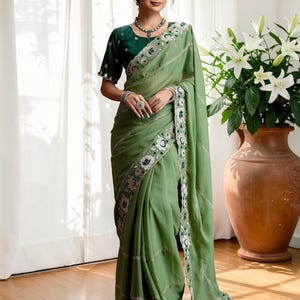 May include: A sage green saree featuring a dark green blouse with short sleeves. The saree is adorned with a wide, decorative border showcasing white floral embroidery and silver accents. The fabric drapes elegantly, complemented by a matching necklace.