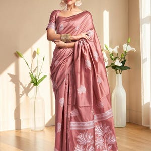 Puede incluir: Un sari de color rosa empolvado con un acabado suave y satinado y bordados florales blancos. La prenda incluye una blusa a juego de manga corta, intrincados patrones de flores de loto en el borde y una silueta drapeada con pliegues elegantes.