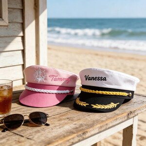 May include: Two captain hats, one pink with a white anchor emblem and the name Tammy embroidered in pink, and one white with a black brim, gold braided trim, and the name Vanessa embroidered in black, both featuring white or gold rope accents.