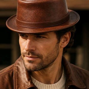 May include: A brown leather pork pie hat with a flat crown and a narrow, upturned brim. The hat features a smooth, consistent texture and a matching leather band encircling the base of the crown, providing a structured and classic appearance.