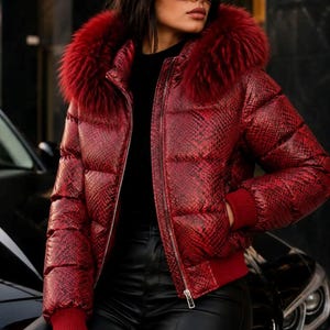 Könnte beinhalten: Eine rote Steppjacke mit Schlangenledermuster, Frontreißverschluss und einer Kapuze mit dickem, rotem Kunstfellbesatz. Die Jacke verfügt über gerippte rote Bündchen an den Ärmeln und am Saum sowie horizontale Steppnähte für eine körpernahe Passform.
