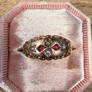 May include: A gold ring featuring an elongated, scalloped-edge setting. The center of the ring is adorned with two small, round, vibrant red stones and three tiny, white, spherical pearls arranged in a cross-like pattern within the metal band.