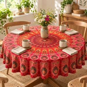 Op de afbeelding: Een ronde tafel bedekt met een rood en oranje tafelkleed met een mandala-patroon. De tafel is gedekt met vier beige keramische mokken op gevlochten onderzetters, vier gevouwen beige servetten en een centrale vaas met kleurrijke veldbloemen.
