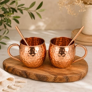 May include: Two round copper mugs with a hammered texture, each featuring a curved handle and a matching copper straw. The mugs have a polished metallic finish and are positioned side by side, reflecting light across their rounded surfaces.