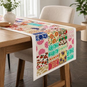 Op de afbeelding: Een rechthoekige tafelloper met een patchworkontwerp van diverse bloem- en geometrische patronen in levendige kleuren zoals roze, turquoise, oranje, blauw en goud, afgewerkt met een effen witte rand langs de zijkanten van de stof.
