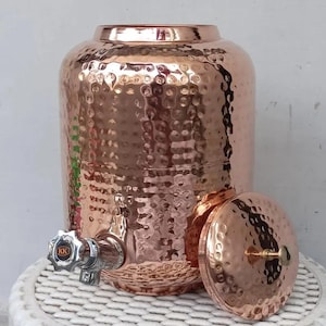 May include: A cylindrical copper water dispenser featuring a hammered texture across its surface. It includes a metallic spigot with a black knob marked with the letters KK and a matching circular lid with a small central handle knob.