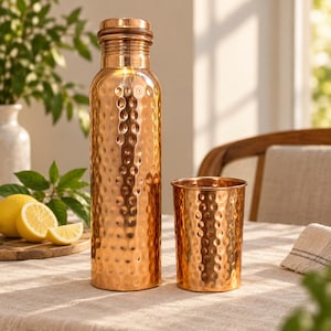 Peut inclure: Une grande bouteille d'eau cylindrique en cuivre avec une texture martelée et un bouchon à vis assorti, accompagnée d'un gobelet en cuivre martelé plus petit. Les deux objets présentent une finition métallique polie aux tons brun-rouge.