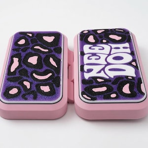 May include: A rectangular pink plastic case with two hinged sections. The top surfaces feature a purple glittery leopard print pattern. The right section displays the white text "NEE DOH" in a stylized font, while the left section shows the pattern.