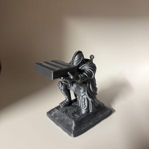 Może przedstawiać: Ciemnoszara, monochromatyczna figurka rycerza w pełnej zbroi płytowej, klęczącego na prostokątnej podstawie. Rycerz jest przedstawiony w pochylonej do przodu postawie, trzymając przed sobą prostokątny blok z trzema poziomymi rowkami na górze.