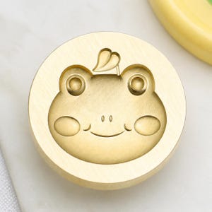 May include: A circular brass wax seal stamp featuring an engraved frog face design. The frog has two prominent eyes, a small nose, a curved mouth, and two oval cheeks. A single leaf shape is positioned directly above the frog's head.