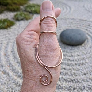 May include: A rose gold-colored metal finger splint featuring a thin band at the top and a larger, curved wire structure that wraps around the base of the thumb, terminating in a decorative spiral design at the bottom of the thumb joint.