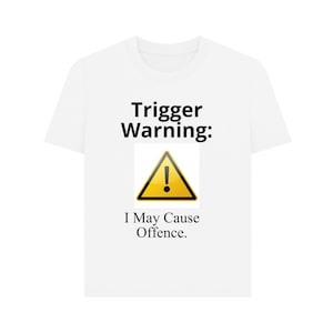 Puede incluir: Una camiseta blanca de manga corta con el texto negro Trigger Warning: en la parte superior, un símbolo de peligro triangular amarillo con un signo de exclamación negro en el centro y el texto negro I May Cause Offence. debajo del símbolo.