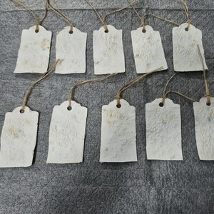 May include: Ten rectangular off-white paper tags with a textured, fibrous surface. Each tag features a rounded top with a punched hole and a thin brown twine loop attached for hanging. The tags are arranged in two rows of five on a dark gray surface.