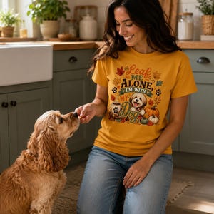 Könnte beinhalten: Ein senfgelbes Kurzarm-T-Shirt mit einem Grafikdruck von einem Hund, Kürbissen, Herbstblättern und einer Kaffeetasse. Der Text lautet Leaf ME ALONE, I'M WITH MY Dog. Das Design enthält Pfotenabdrücke und herbstliches Laub in Orange, Braun und Grün.