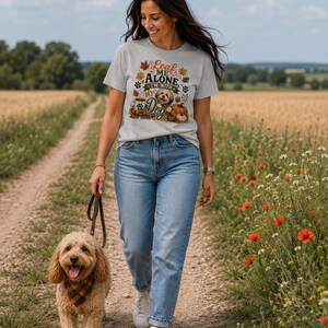 Könnte beinhalten: Ein hellgraues Kurzarm-T-Shirt mit einem Grafikdruck aus Herbstblättern, Kürbissen, einer Kaffeetasse und einem Hund sowie dem Text "Leaf ME ALONE I'M WITH MY Dog". Das T-Shirt wird mit einer hellblauen Jeans und weißen Turnschuhen kombiniert.