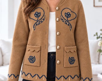 Puede incluir: Una chaqueta de color tostado con cuello redondo, mangas largas y cinco botones dorados. La prenda está decorada con motivos florales bordados en azul oscuro en el pecho y los bolsillos, y un patrón geométrico en zigzag en el dobladillo.