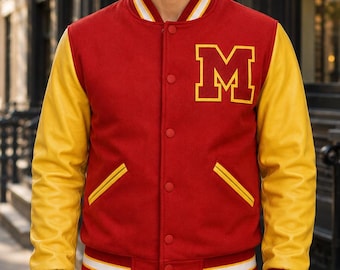 Puede incluir: Una chaqueta universitaria roja con mangas amarillas, que presenta una letra M grande de color amarillo en el pecho izquierdo. La chaqueta tiene cierre frontal con botones a presión, dos bolsillos laterales con ribete amarillo y puños y cuello acanalados.