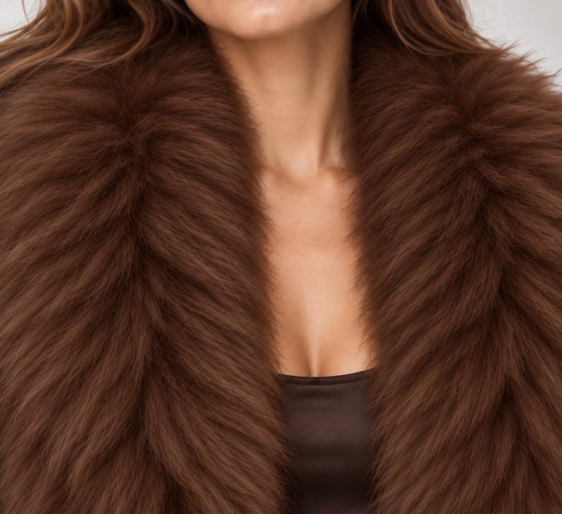 May include: A thick, plush brown faux fur collar draped around the neck and shoulders. The collar features long, dense, textured fibers in a deep chocolate brown shade, creating a voluminous and layered appearance over a dark brown top.