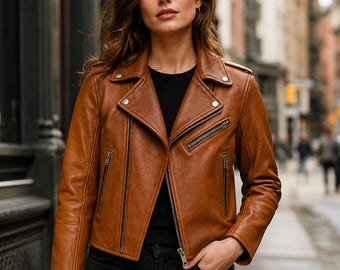 Puede incluir: Una chaqueta de cuero color coñac estilo motero con solapa de muesca, cremalleras metálicas plateadas en la parte delantera y el bolsillo del pecho, y detalles de botones a presión en el cuello. Se lleva sobre una camiseta negra y vaqueros negros.