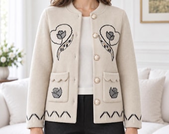 Puede incluir: Una chaqueta beige de manga larga con cuello redondo, cinco botones redondos nacarados y dos bolsillos delanteros con bordes festoneados. La chaqueta está decorada con motivos florales bordados en gris oscuro y un patrón geométrico en el dobladillo.