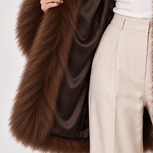 May include: A brown faux fur coat with a smooth dark brown satin lining, worn over a white top and light beige high-waisted trousers. The coat features long, dense brown faux fur trim along the front opening and a structured, tailored fit.