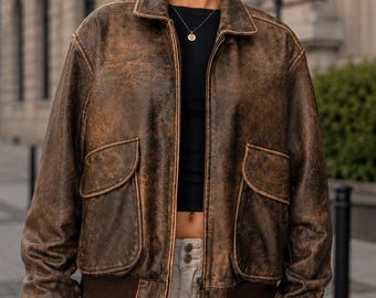 Puede incluir: Una chaqueta tipo bomber de cuero marrón desgastado con cuello clásico, cierre de cremallera frontal y dos grandes bolsillos con solapa. La chaqueta presenta puños de canalé marrón oscuro y un dobladillo a juego con textura envejecida.