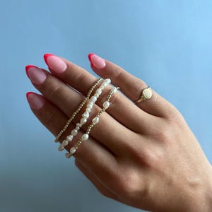 May include: Three delicate bracelets featuring a combination of small metallic gold beads and irregular white freshwater pearls, worn alongside a simple gold signet ring with a textured circular face on the ring finger.