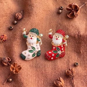 May include: Two Santa Claus brooches, one with a green hat and white stocking, the other with a red hat and red stocking. Both feature gold-tone metal, clear rhinestones, and green enamel holly leaves. The figures have white beards and red lips.