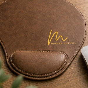 May include: A brown leather mouse pad with a wrist rest, featuring a yellow cursive letter M and the text MORGAN MAXWELL printed in yellow. The mouse pad has visible stitching along the edges and a smooth, textured surface for computer use.