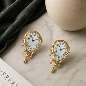 May include: A pair of gold-toned earrings shaped like melting clocks. Each earring features a white clock face with black numerals and black hands, surrounded by a fluid, dripping gold metal frame that mimics a distorted, liquid appearance.