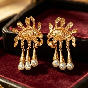 May include: A pair of gold-toned earrings featuring an eye motif with a textured, irregular sunburst-like border. Each earring has three dangling gold-toned cone-shaped charms, each tipped with a small, round, white pearl-like bead.