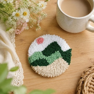 May include: A circular punch needle coaster featuring a landscape design with a small pink sun, dark green and light green mountain peaks, and a textured beige foreground. The coaster is crafted with thick yarn loops in a dense, tactile pattern.