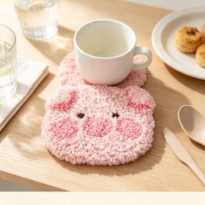 May include: A pink pig-shaped punch needle coaster with a textured, looped yarn surface, featuring dark brown embroidered eyes and a stitched snout, supporting a plain white ceramic mug on a light wooden surface with the text MossPunchCraft.