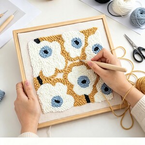 May include: A square wooden embroidery hoop containing a punch needle project on white fabric. The design features stylised flowers with white petals, light blue centres, and mustard yellow backgrounds. A wooden punch needle tool is in use.