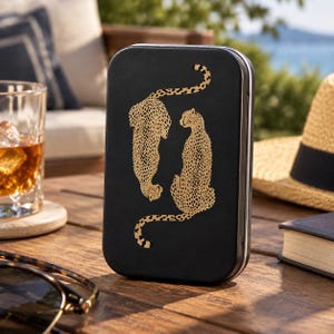 May include: A rectangular black metal tin featuring a gold-colored graphic of two cheetahs. One cheetah is positioned facing downward while the other sits upright, both displaying detailed spotted patterns against the matte black surface of the container.