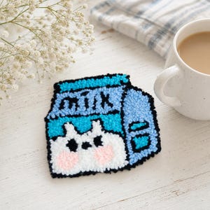 May include: A punch needle coaster shaped like a milk carton in shades of blue, white, and black. The design features the word milk in black lettering above a white cat face with pink cheeks. The coaster has a thick, textured yarn finish.