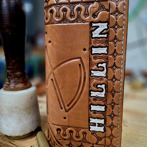 May include: A tan leather wallet featuring intricate hand-tooled geometric and star patterns along the border. The wallet displays the name HILLIN in white lettering vertically on the right side, with a large embossed arch design on the left.
