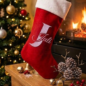 Könnte beinhalten: Ein roter Weihnachtsstrumpf aus Samt mit einem weißen Kunstfell-Umschlag. Der Strumpf ist mit einem großen, glitzernden silbernen Buchstaben J verziert, über den der Name Juli in einer kursiven rosa Schrift geschrieben ist. Er hängt an einer Schlaufe.