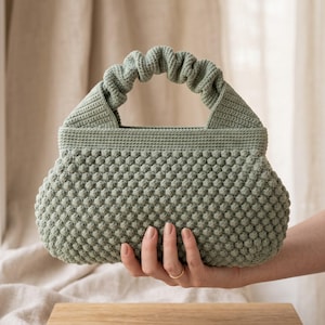May include: A sage green crochet handbag featuring a textured bobble stitch pattern across the body. The bag has a structured rectangular opening and a single gathered, ruched handle attached to the top, crafted from the same sage green yarn.