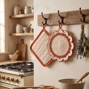 May include: Two crocheted pot holders hanging on a wooden wall rack. One is square with vertical cream stripes and a rust-coloured border. The other is circular with a spiral cream centre, a rust-coloured ring, and a scalloped cream edge.