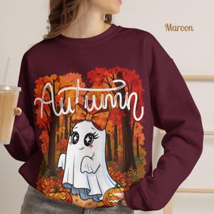 Puede incluir: Una sudadera granate de manga larga con un estampado gráfico de un fantasma blanco con un lazo de cuadros naranja. El diseño incluye la palabra Autumn en letra cursiva blanca, un fondo de bosque con follaje otoñal naranja y dos calabazas.