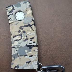 May include: A curved, rectangular magazine-shaped keychain featuring a multi-colored camouflage pattern in shades of tan, gray, black, and white. It includes a circular silver-toned metal accent on the top and a dark metal carabiner attached to the base.